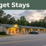 A roadside motel entrance beside a car, pine trees, and a quiet highway.