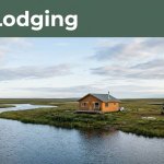 A lit cabin beside a river, tundra, and parked bush plane.