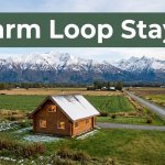 A cozy cabin beside farmland with snowy mountains and a gravel road.