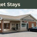 Budget motel exterior with a parked car, lake scenery, and a Budget Stays banner.