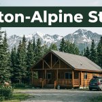 A rustic cabin lodge and SUV sit beside an Alaska highway beneath snowy mountains.
