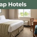 A tidy hotel room with a made bed, small desk, suitcase, and window view.
