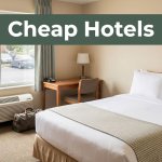 Budget hotel room with a made bed, small desk, travel bag, and parking lot outside.