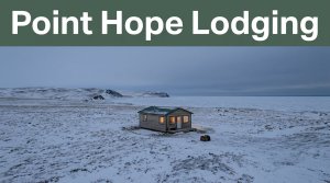 A warm-lit cabin stands in snowy Point Hope tundra beside a travel bag.