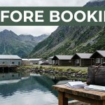 A Sand Point motel and harbor sit below Aleutian mountains, with a travel bag and booking papers in front.