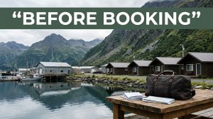 A Sand Point motel and harbor sit below Aleutian mountains, with a travel bag and booking papers in front.