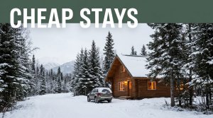 Wood cabin with warm windows beside a snowy road, spruce trees, mountains, and a parked vehicle.