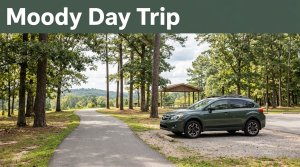 A parked car beside a paved trail in a sunny Alabama park with trees, a pavilion, and distant hills.
