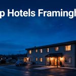 A modest Framingham hotel glows at blue hour beside a parked car and distant highway lights.
