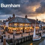 A riverside inn, historic street, quay, and sailboats at sunset.