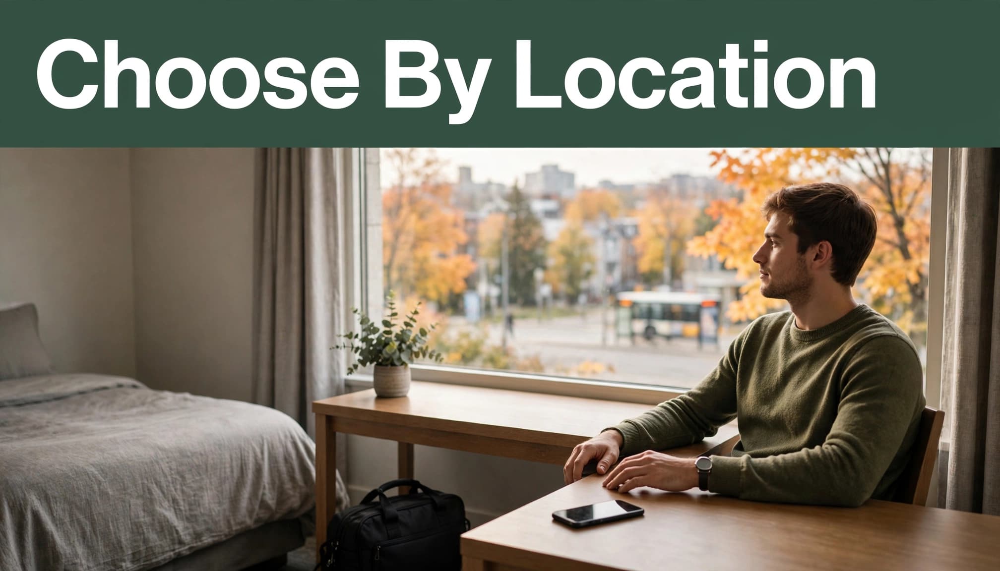 Traveler seated at a hotel desk beside a bag and phone, with autumn trees outside.
