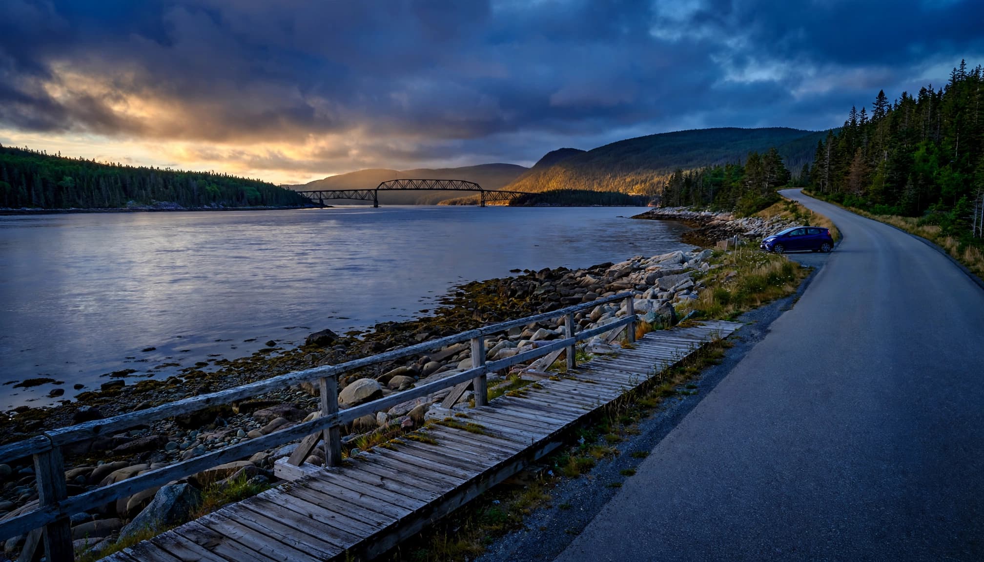 A coastal road and boardwalk beside calm water, forested hills, and a distant bridge.